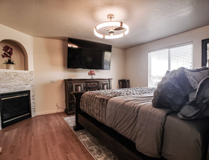 Bedroom with dark wood furniture, stone fireplace, wall-mounted TV, ceiling fan light and natural light from window