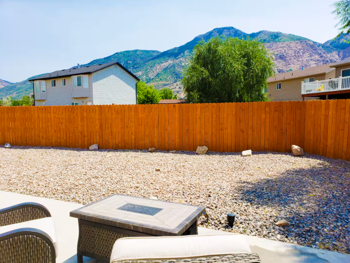 Large fenced backyard with gravel landscaping, fire pit seating, mature willow tree and dramatic Wasatch Mountain backdrop