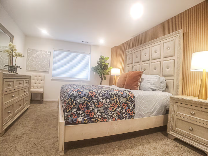 Spacious master bedroom with tall tufted panel headboard, floral bedding, matching dressers, warm lamps and fiddle leaf fig tree