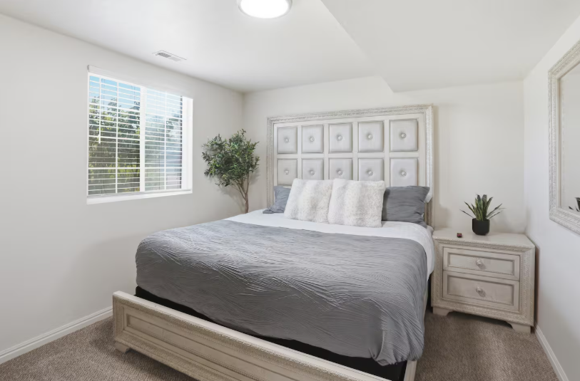 King bedroom with tall white tufted panel headboard, gray bedding, faux olive tree, nightstand with plants and bright window