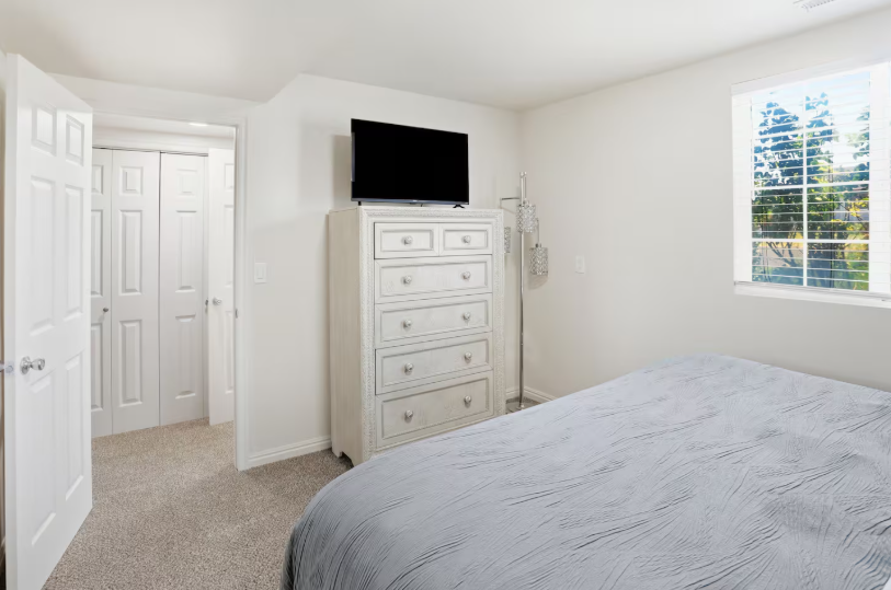 Bedroom with wall-mounted TV on white tall dresser, gray bedding, double closet doors and bright window with tree view