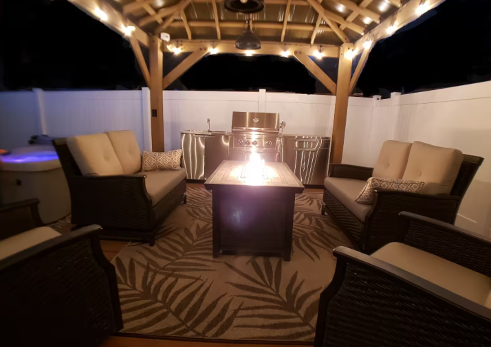 Backyard gazebo at night with string lights, fire pit table, wicker lounge seating, outdoor kitchen with BBQ grill and hot tub glowing in background