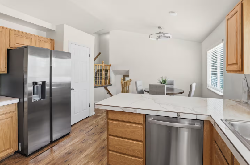 Kitchen with honey oak cabinets, marble island, stainless steel refrigerator and dishwasher, hardwood floors and open view to dining nook and staircase