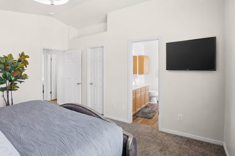 Master bedroom with tufted gray velvet bed, wall-mounted TV, vaulted ceiling, faux tree and open door to ensuite bathroom